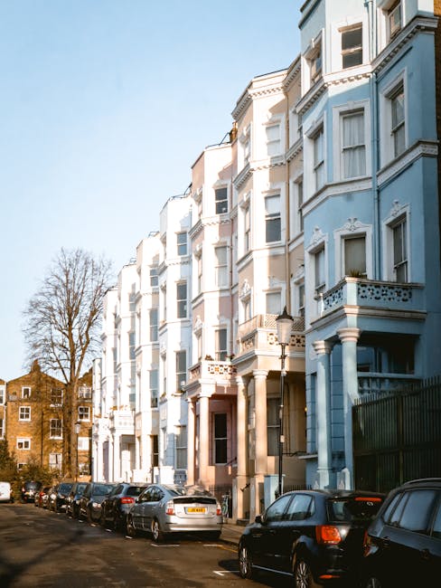 Photograph of a multi-storey residential building with a light blue facade, featuring ornate white window frames and decorative balcony railings, set against a clear blue sky. The street in front of t