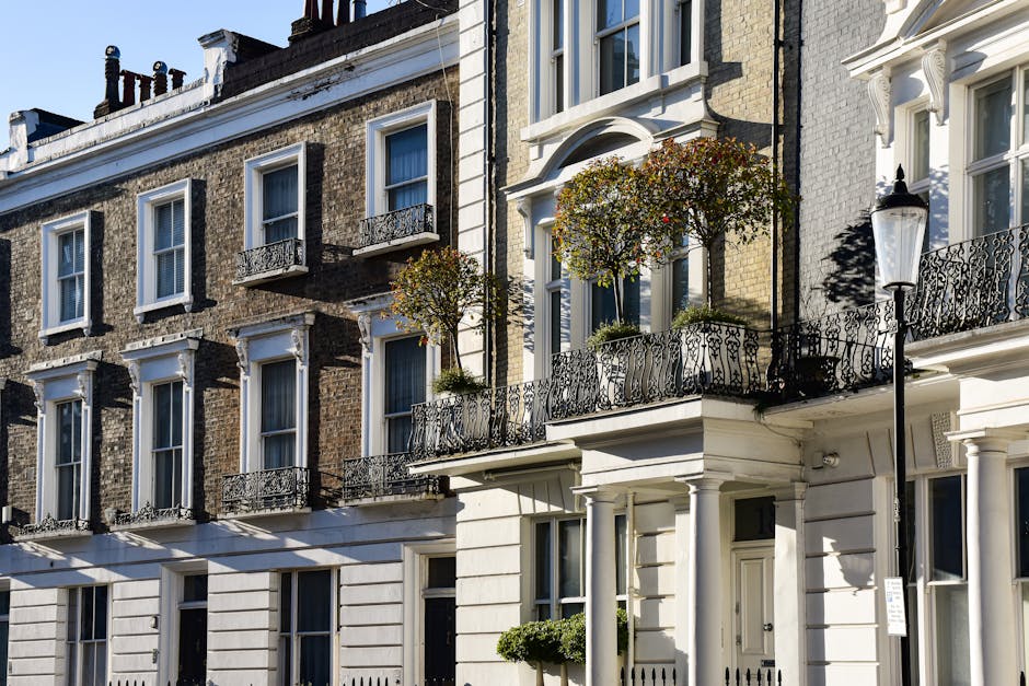 Photograph of a multi-storey residential building with a light blue facade, featuring ornate white window frames and decorative balcony railings, set against a clear blue sky. The street in front of t