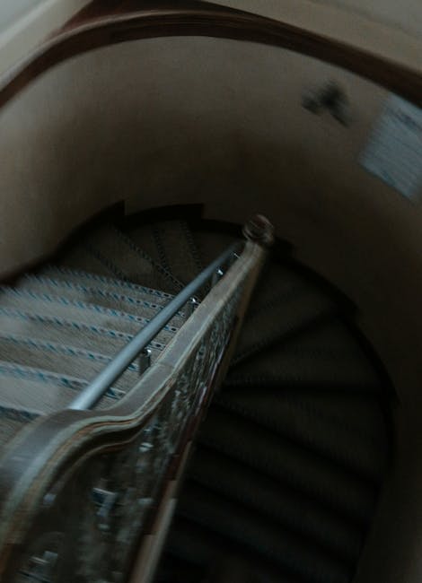 A spiral staircase inside a residential property with carpeted steps and a decorative wooden and metal handrail. The staircase curves downward from an upper floor, with the wall on the inner side painted in a light beige color. On the outer side, a small section of a window with blinds is visible, allowing natural light into the space. The photo captures a home relocation scenario, with the focus on the staircase as a narrow passage for moving large furniture or boxes. The environment appears clean and well-maintained, consistent with professional removals involving cautious navigation of tight staircases, as facilitated by Removal Company Pimlico.