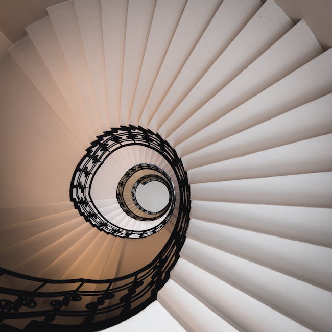 A downward view of a spiral staircase inside a building, featuring white steps with a smooth finish and a black wrought iron railing following the curve of the staircase. The staircase appears to descend through multiple levels, with a round central opening visible at the bottom. The lighting is soft, ensuring the staircase is well-illuminated without harsh shadows. This interior scene is typical of a residential property undergoing a home relocation, with the staircase ready for the movement or packing of furniture and boxes by professionals such as Removal Company Pimlico, specializing in house removals and transport logistics.
