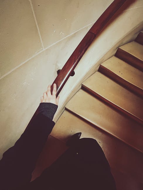 A spiral staircase inside a residential property with carpeted steps and a decorative wooden and metal handrail. The staircase curves downward from an upper floor, with the wall on the inner side painted in a light beige color. On the outer side, a small section of a window with blinds is visible, allowing natural light into the space. The photo captures a home relocation scenario, with the focus on the staircase as a narrow passage for moving large furniture or boxes. The environment appears clean and well-maintained, consistent with professional removals involving cautious navigation of tight staircases, as facilitated by Removal Company Pimlico.