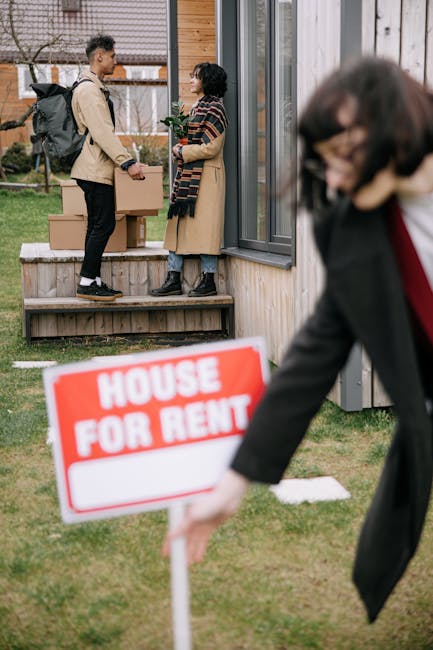 A red and white 'House For Rent' sign is positioned on a grassy area outside a property, with a wooden fence in the background. Behind the sign, three individuals are standing nearby, engaged in conve